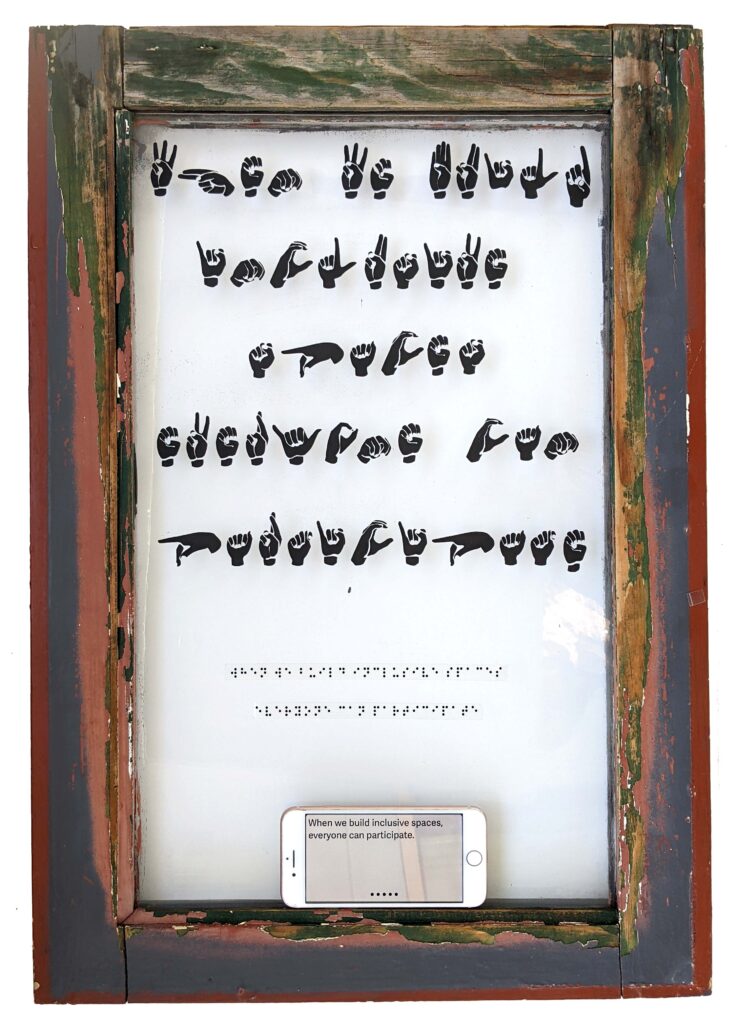 Artwork on the glass of an old window. Decals, or stickers, of hands fingerspelling, and underneath, Braille spells out the message of the piece. A phone sits at the bottom of the window, providing live captioning. The fingerspelling, Braille, and phone say "When we build inclusive spaces, everyone can participate." By Diana Kohne