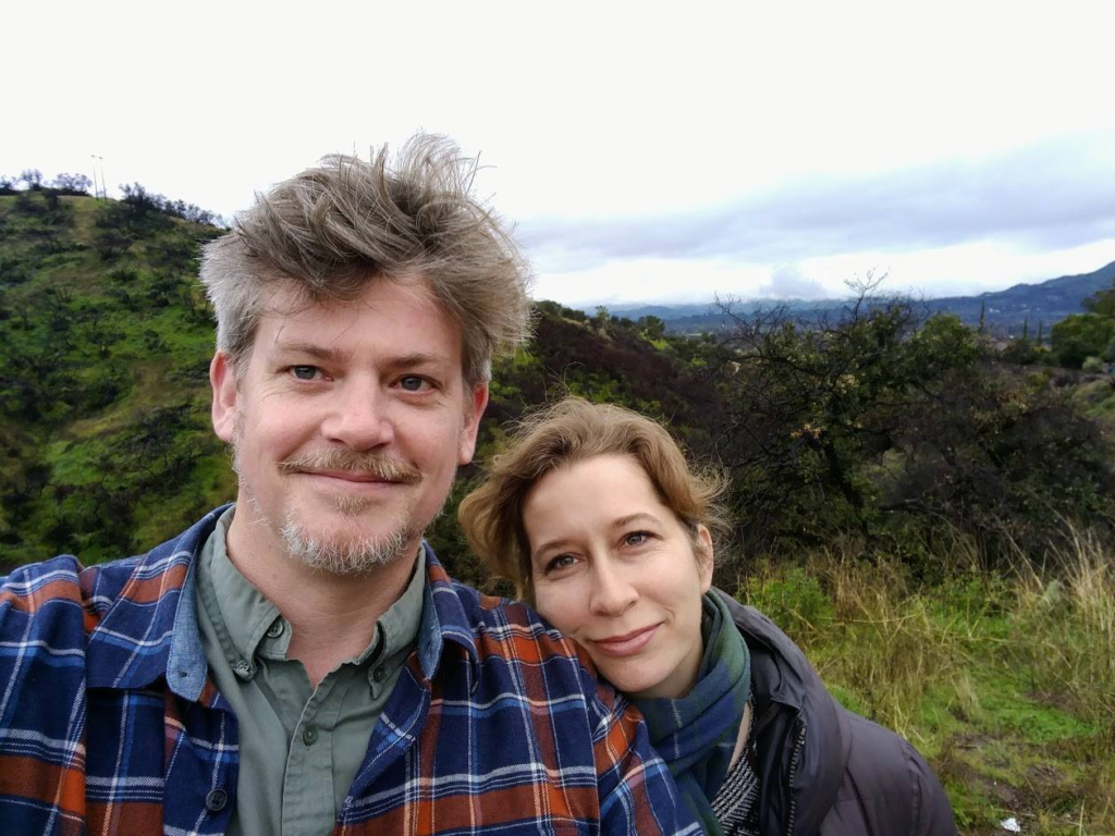 photo of Altadena native garden designers Michael and Diana. Michael has grey hair and beard and a plaid shirt and diana has dark blond hair in a pony tail. She wears a plaid scarf as they stand in front of a sweeping California native background